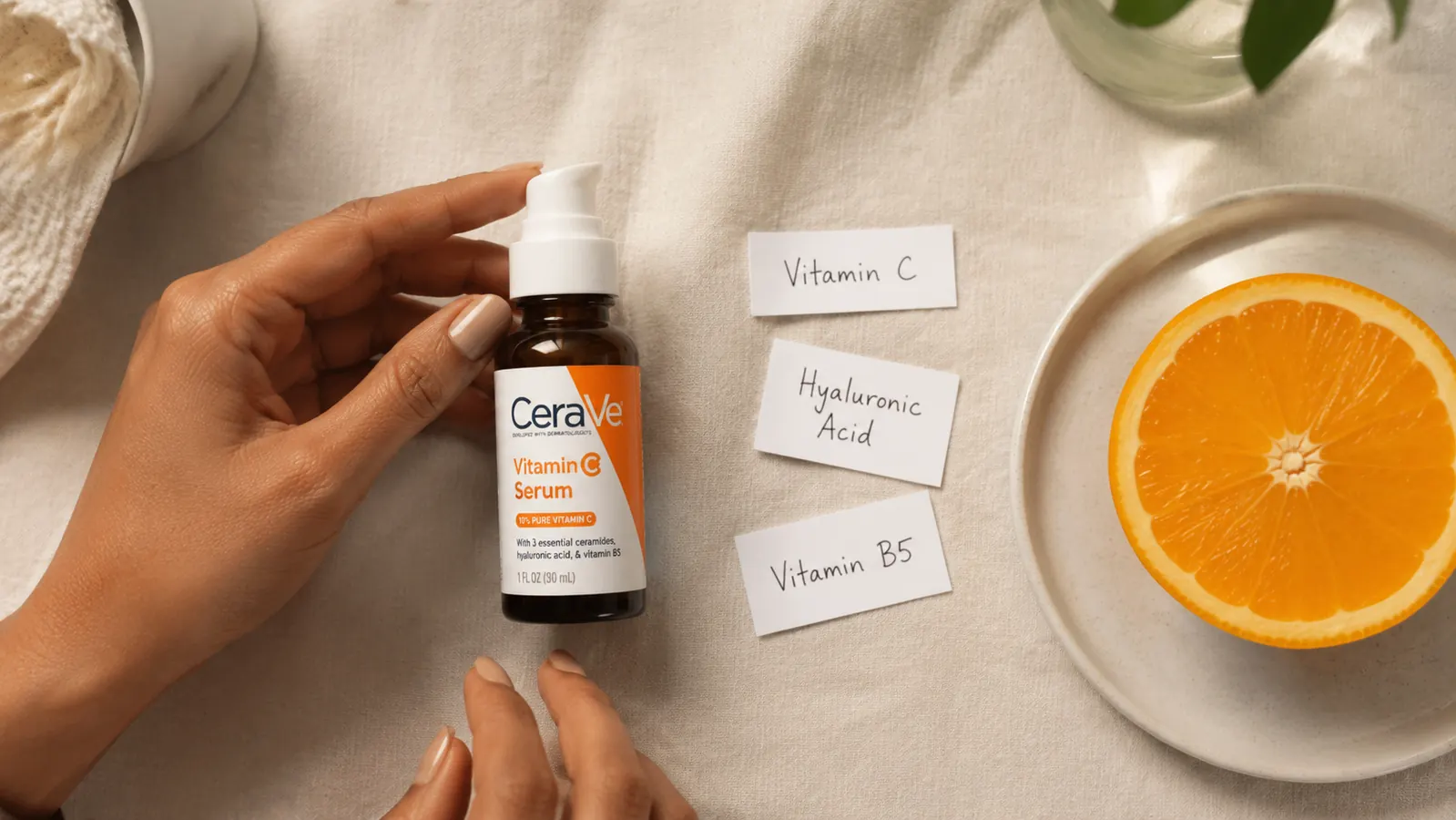 Flat lay of CeraVe Vitamin C Serum bottle next to a vitamin C orange slice and a few ceramide ingredient cards on a light cream background