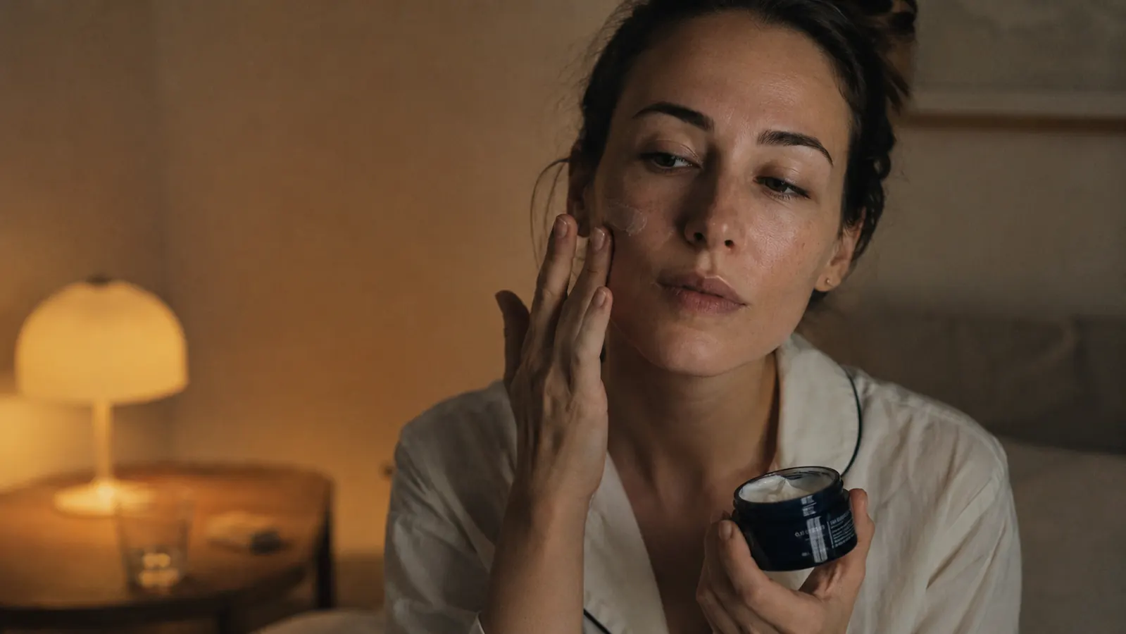A person applying face cream gently at night with soft bedside lamp light, calm and routine-feeling