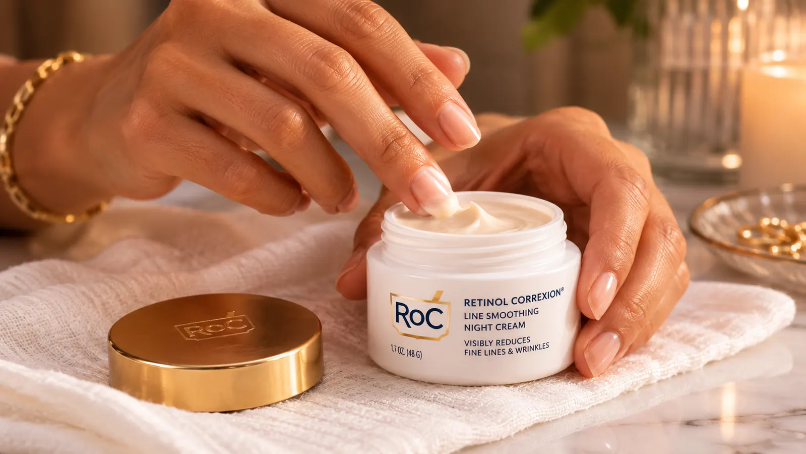 Hand applying a small amount of night cream from a jar onto fingertips over a white towel